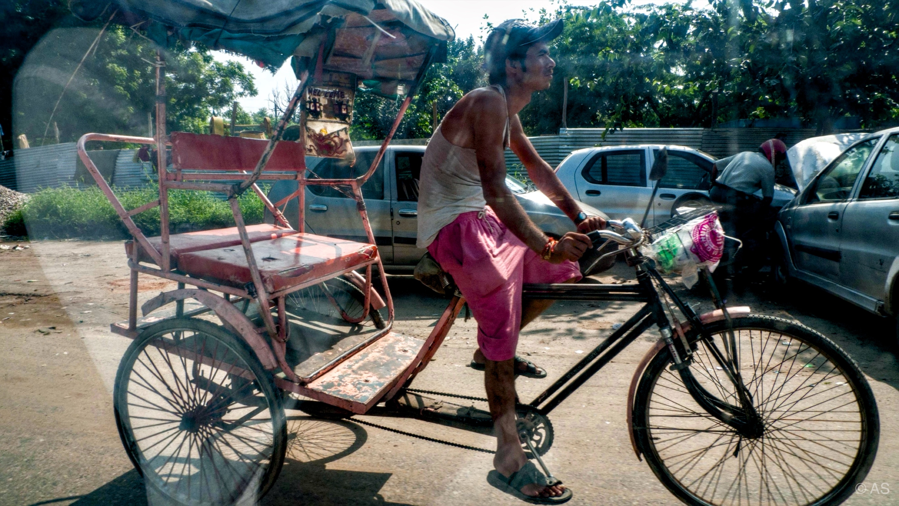 Rickshaw driver cycling past parked cars on a dusty Indian street