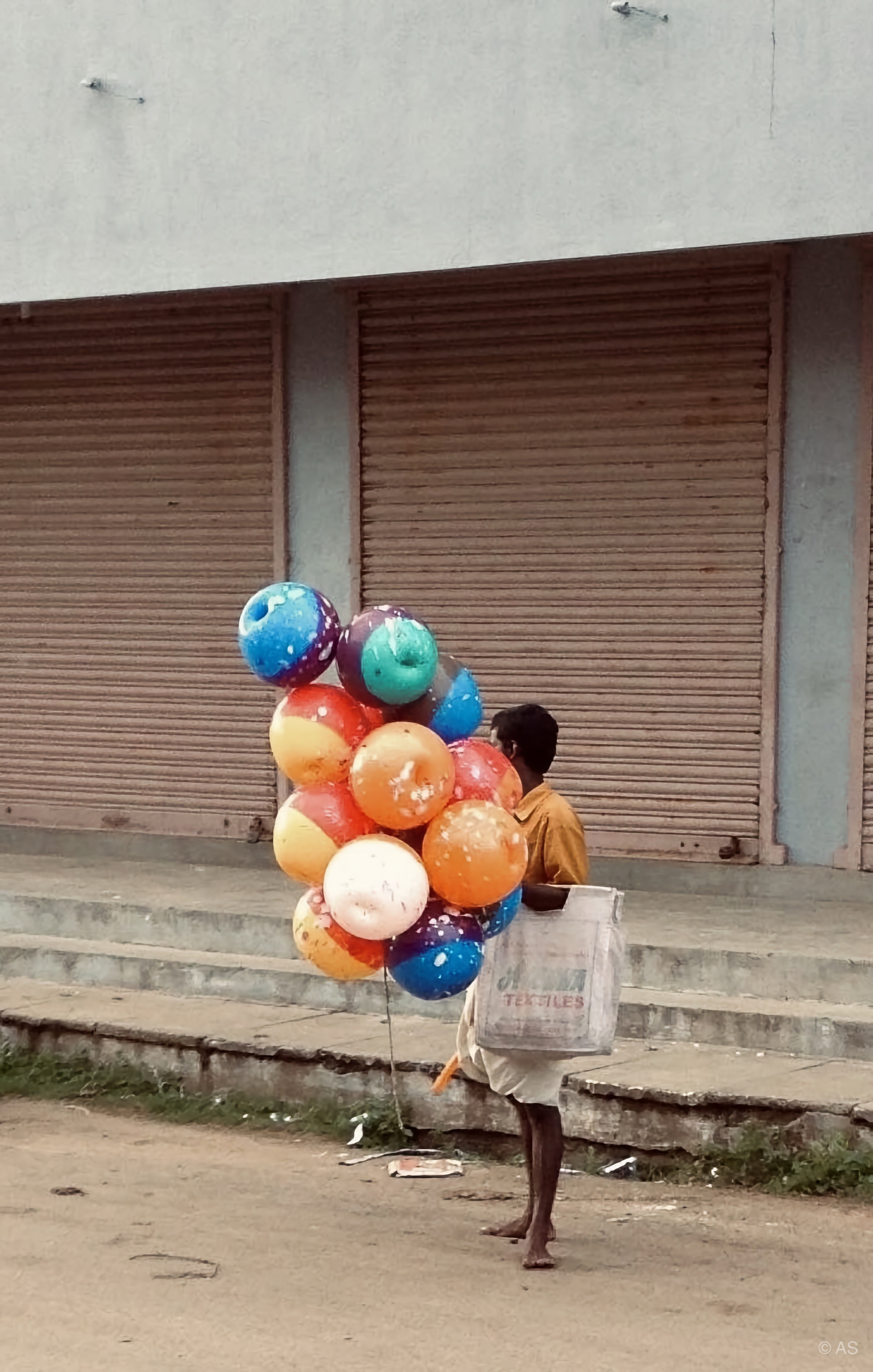 Balloon seller walking past closed metal shutters with a bright cluster of balloons