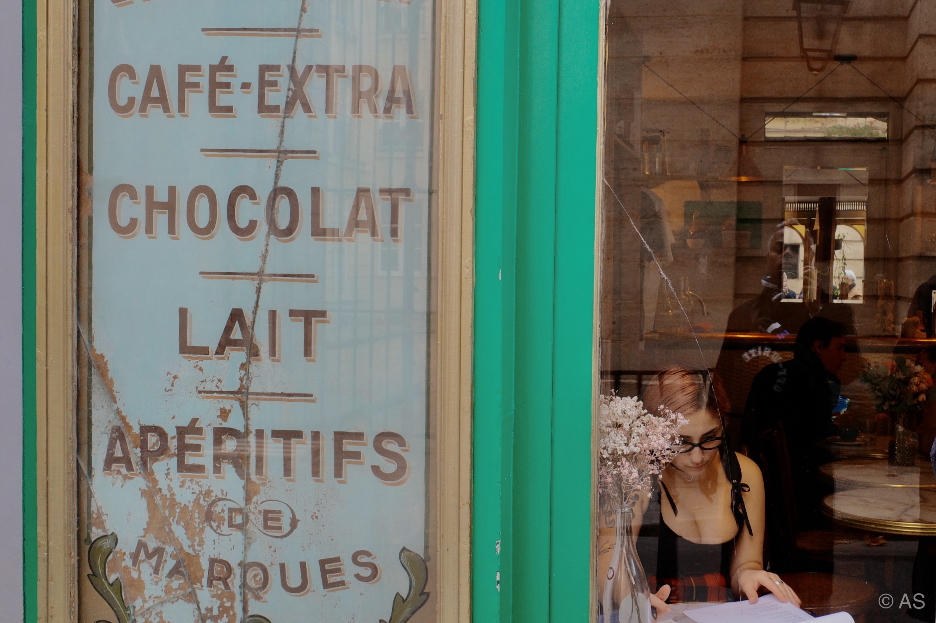 Vintage Parisian café window with painted menu lettering and a woman reading inside